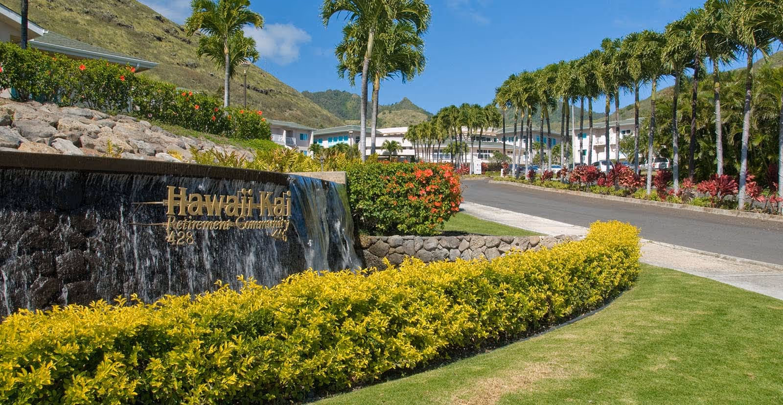 Entrance to Hawaii Kai Retirement Community with a stone wall waterfall feature displaying the community name, surrounded by lush landscaping including yellow and red bushes, palm trees, and a clear blue sky with mountains in the background.