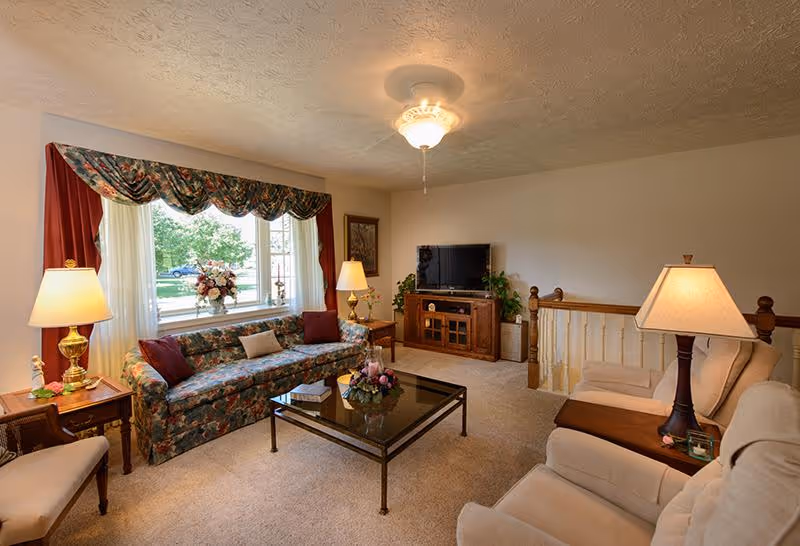 A cozy living room with a floral patterned sofa, two beige armchairs, a glass coffee table with a floral centerpiece, two side tables with lamps, a wooden TV stand with a flat-screen TV, and a large window with floral valance and red curtains letting in natural light.