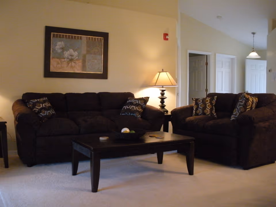 A cozy living room with two dark brown sofas adorned with patterned cushions, a dark wooden coffee table with a decorative bowl and coasters, a side table with a lamp, and a framed floral artwork on the wall. The room has beige walls and carpeted flooring, with doorways visible in the background.