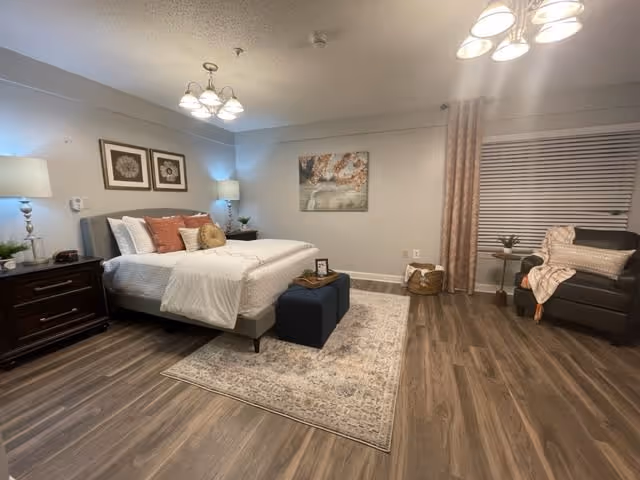 A cozy bedroom with a large bed featuring white bedding and decorative pillows. There are two framed floral artworks above the bed and a nightstand with a lamp and plants on the left side. A patterned rug is placed under the bed, and a small ottoman is at the foot of the bed. On the right side of the room, there is a window with closed blinds and curtains, a small round side table with a plant, and a comfortable armchair with a throw blanket and pillow. The room has wood flooring and soft lighting from ceiling fixtures and lamps.
