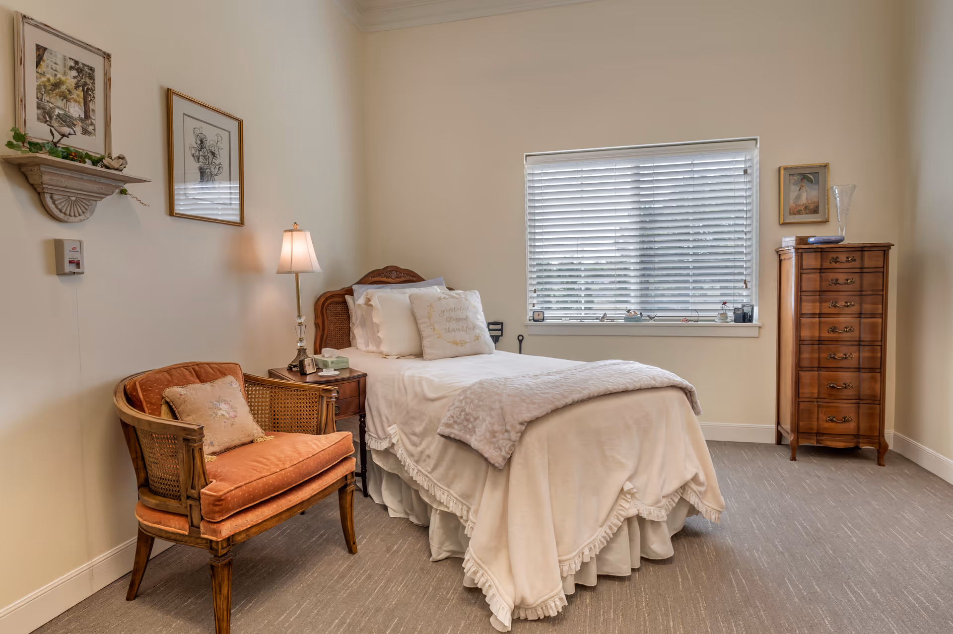 Sunlit single bedroom with a neatly made bed, an upholstered armchair, bedside table with lamp, and a tall wooden dresser.