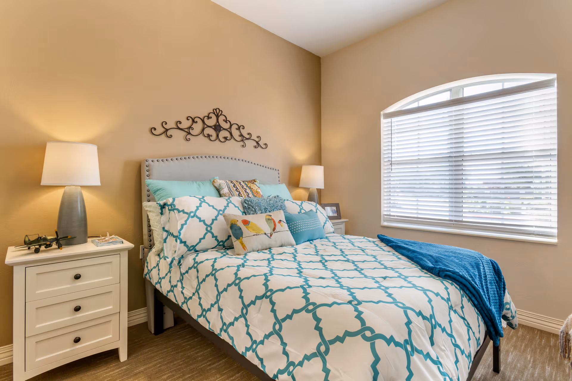 A cozy bedroom with a bed featuring a white and teal patterned comforter and multiple decorative pillows, including one with bird designs. There are two white nightstands on either side of the bed, each with a lamp. A decorative metal wall art piece hangs above the bed. A large window with white blinds allows natural light into the room.