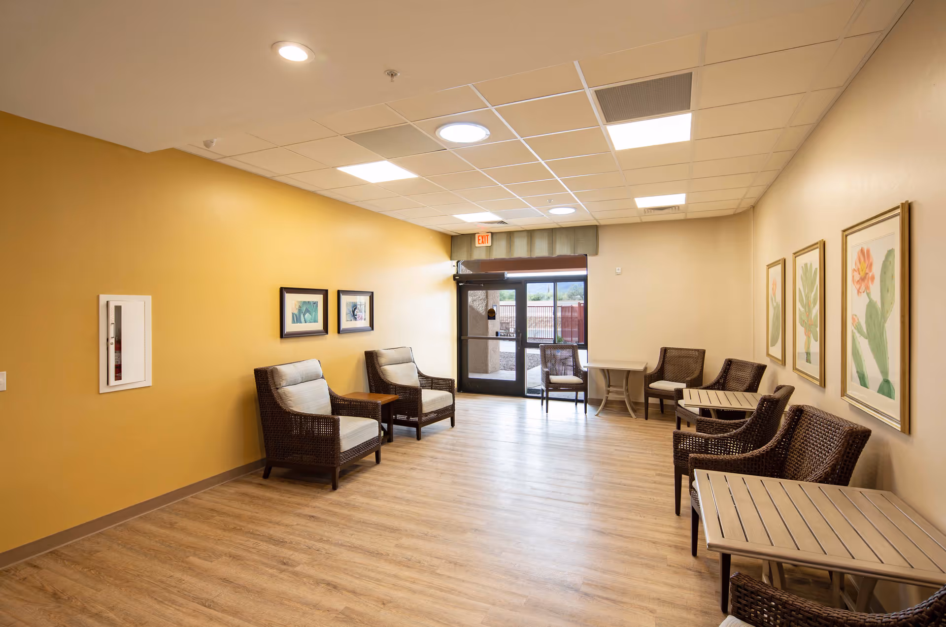 A bright, spacious seating area with wooden flooring and beige walls. The room features several wicker chairs with cushions, small tables, framed botanical artwork on the walls, and a glass door leading outside with a view of a fenced outdoor area and mountains in the distance.