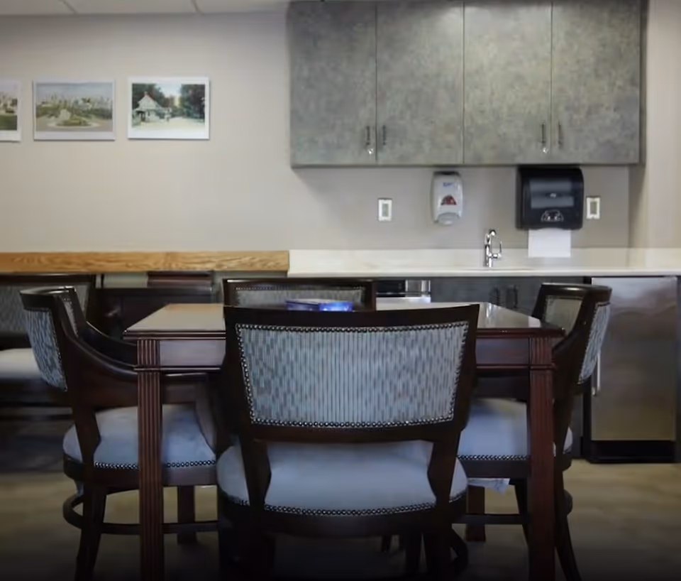 A dining area with a wooden table and four upholstered chairs. Behind the table is a countertop with a sink, soap dispenser, and paper towel dispenser mounted on the wall. Above the countertop are gray cabinets. Three framed pictures hang on the wall to the left.