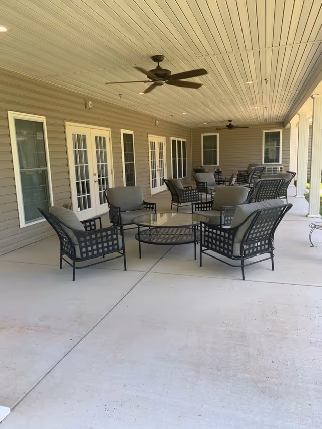 Covered outdoor patio area with multiple cushioned metal chairs and glass-top tables arranged for seating. The patio has a beige ceiling with ceiling fans and recessed lighting, and the building exterior features beige siding with several windows and French doors.