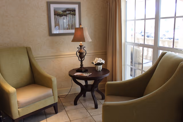 A cozy sitting area with two green upholstered armchairs facing each other, separated by a round wooden side table holding a decorative lamp, a small potted plant, and some reading materials. A framed artwork hangs on the beige patterned wall behind one chair, and large windows with beige curtains let in natural light from outside.