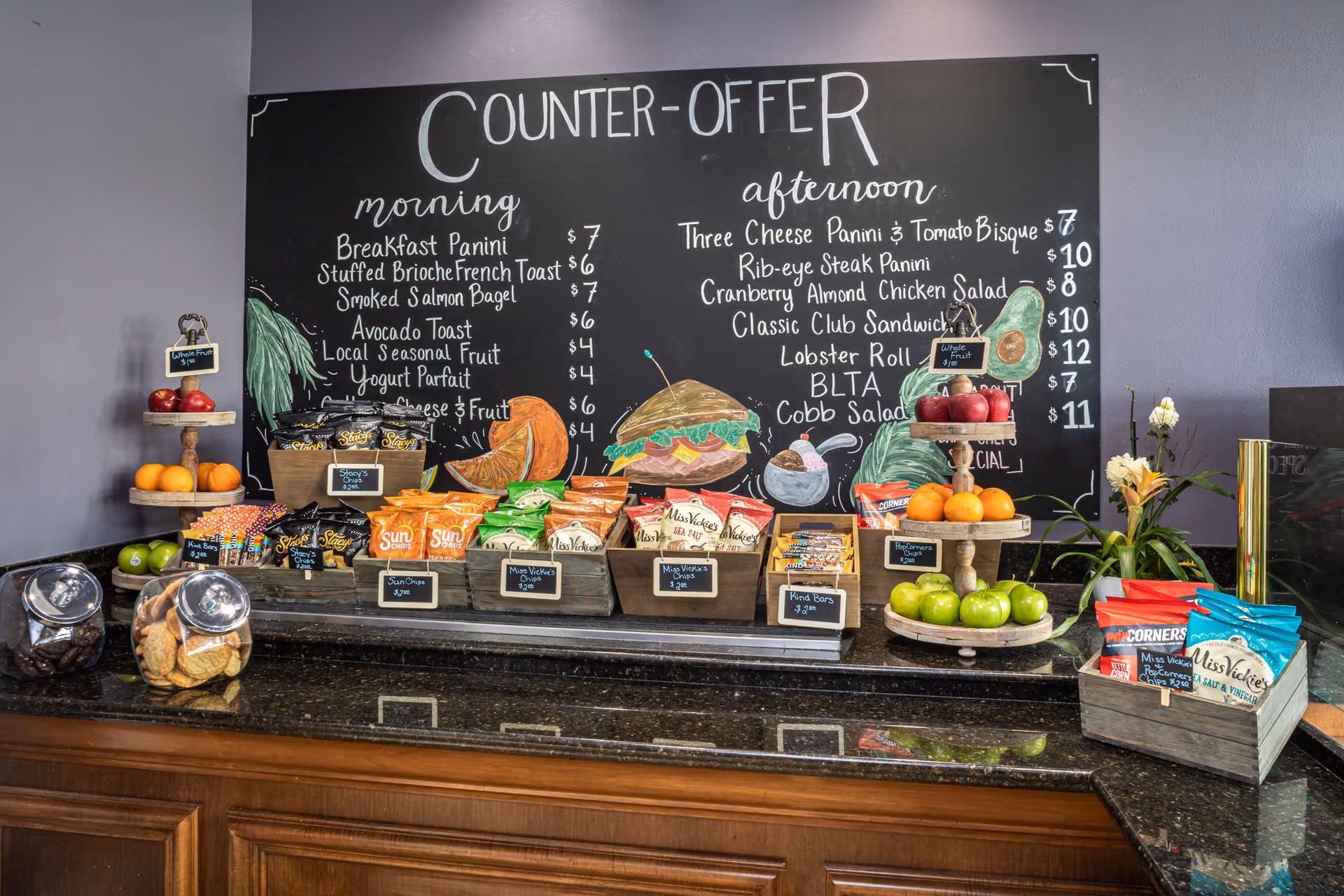 A snack and sandwich counter with a variety of chips, cookies, fruit, and granola bars displayed on a dark granite countertop. Behind the counter is a large blackboard menu listing morning and afternoon food options such as breakfast panini, stuffed brioche french toast, rib-eye steak panini, and lobster roll. The counter is decorated with fresh apples, oranges, and green apples on tiered wooden stands, along with a small plant with yellow flowers on the right side.