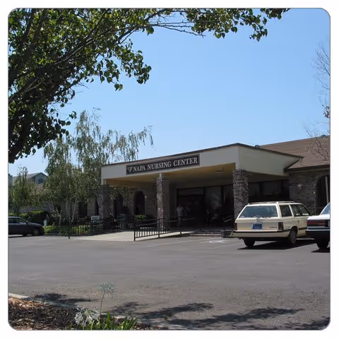 Exterior view of Napa Nursing Center building with a covered entrance supported by stone pillars. Several cars are parked in front of the building under a clear blue sky with some trees nearby.