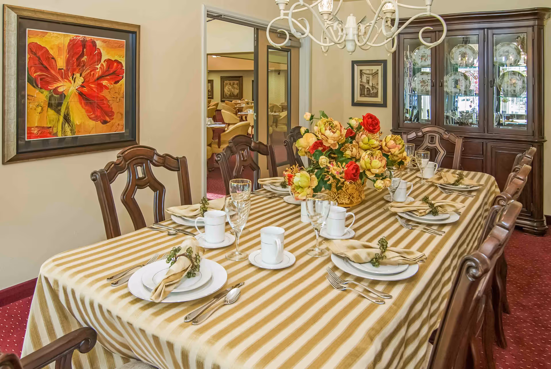 Formal dining room with a long striped tablecloth-covered table set for a meal, a large floral centerpiece, chandelier, wooden chairs and a china cabinet.