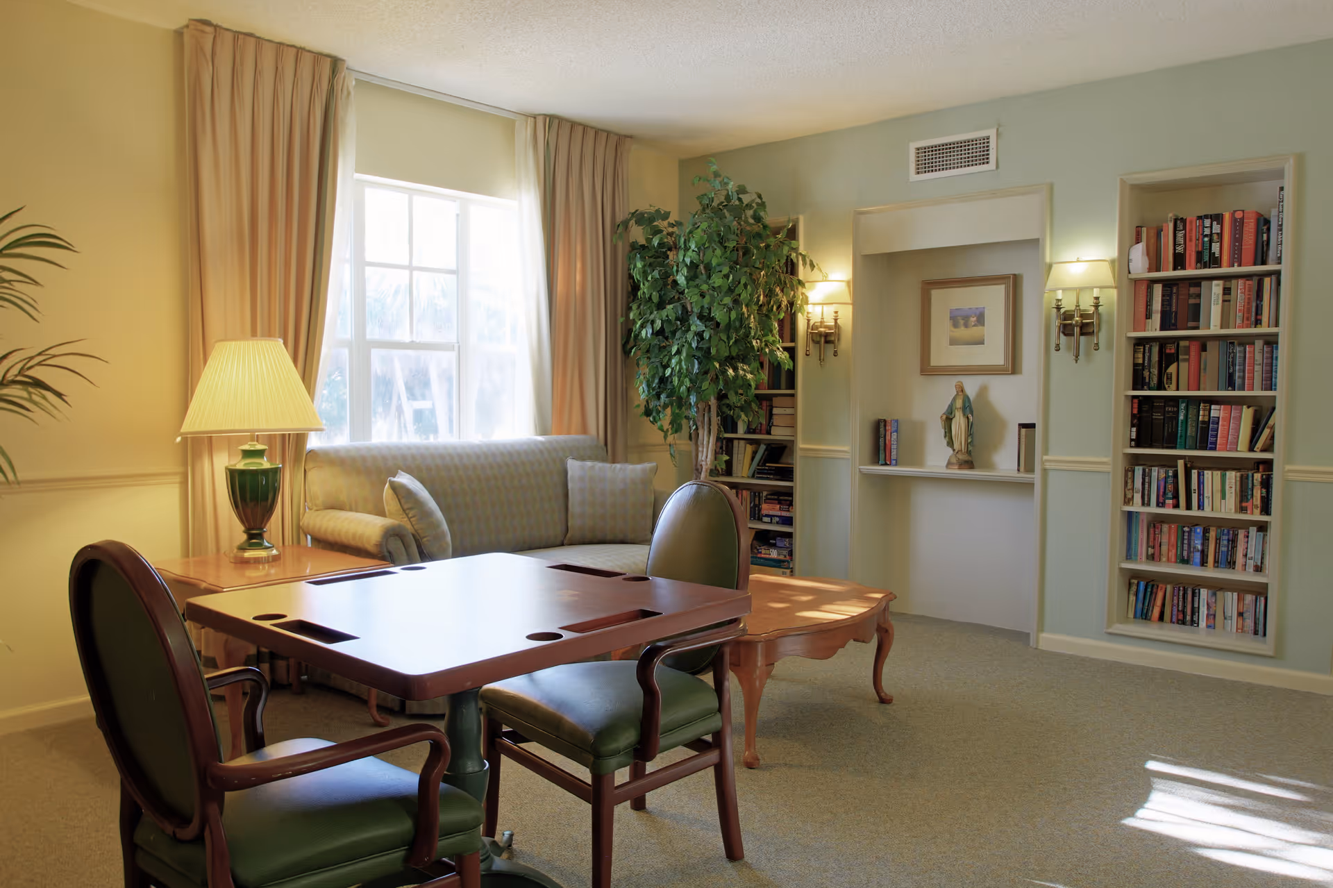A cozy living room with a wooden table and two green cushioned chairs in the foreground, a beige sofa with cushions near a window with beige curtains, a green potted plant, built-in bookshelves filled with books, a small statue, and framed artwork on the wall.