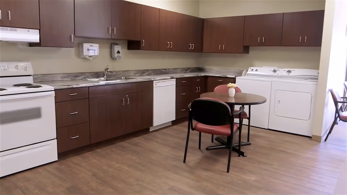 Communal kitchen area with dark wood cabinets, white stove and dishwasher, a washer and dryer, and a small round table with chairs.