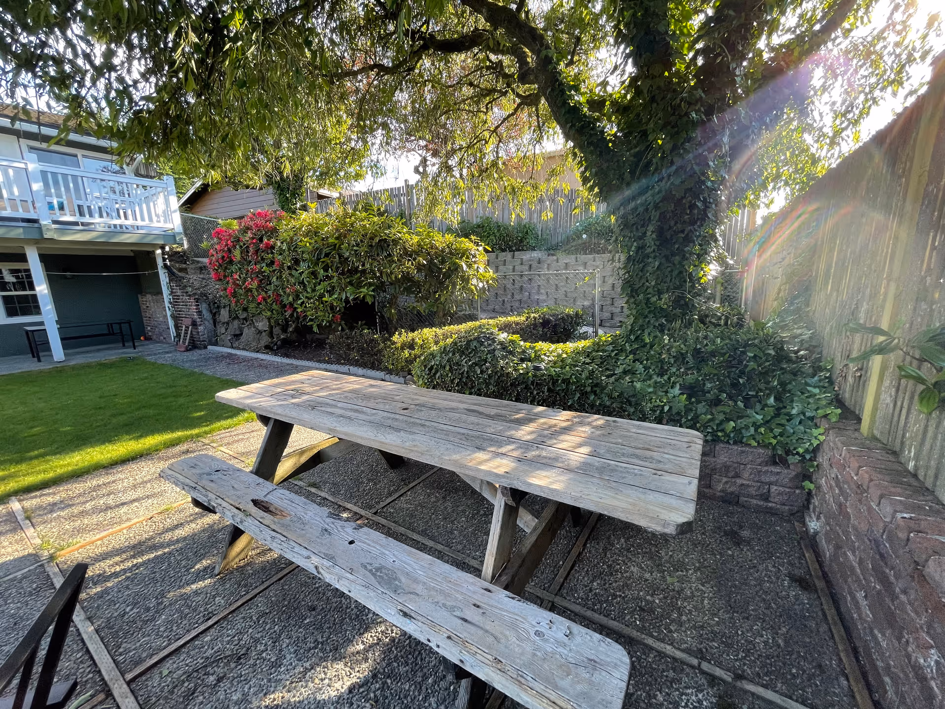 Weathered wooden picnic table and benches on a patio beneath a tree in a landscaped backyard with lawn and a raised deck.