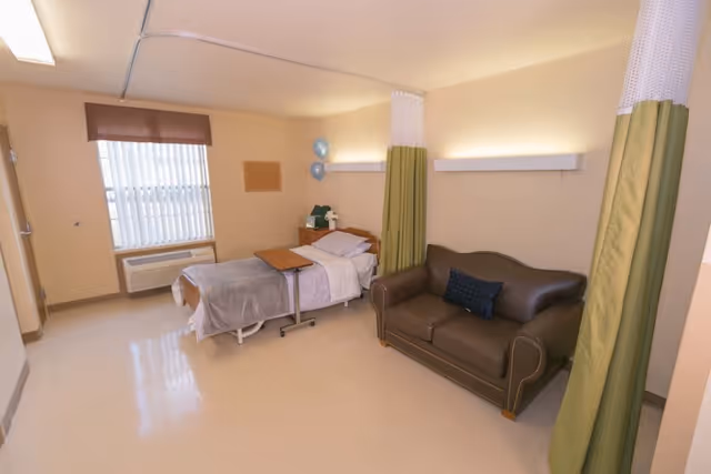 A clean and simple patient room in a healthcare facility featuring a single hospital bed with white and gray bedding, a brown leather loveseat with a dark blue pillow, a small wooden bedside table with a phone and some balloons, a window with vertical blinds and a brown valance, and green privacy curtains partially drawn.
