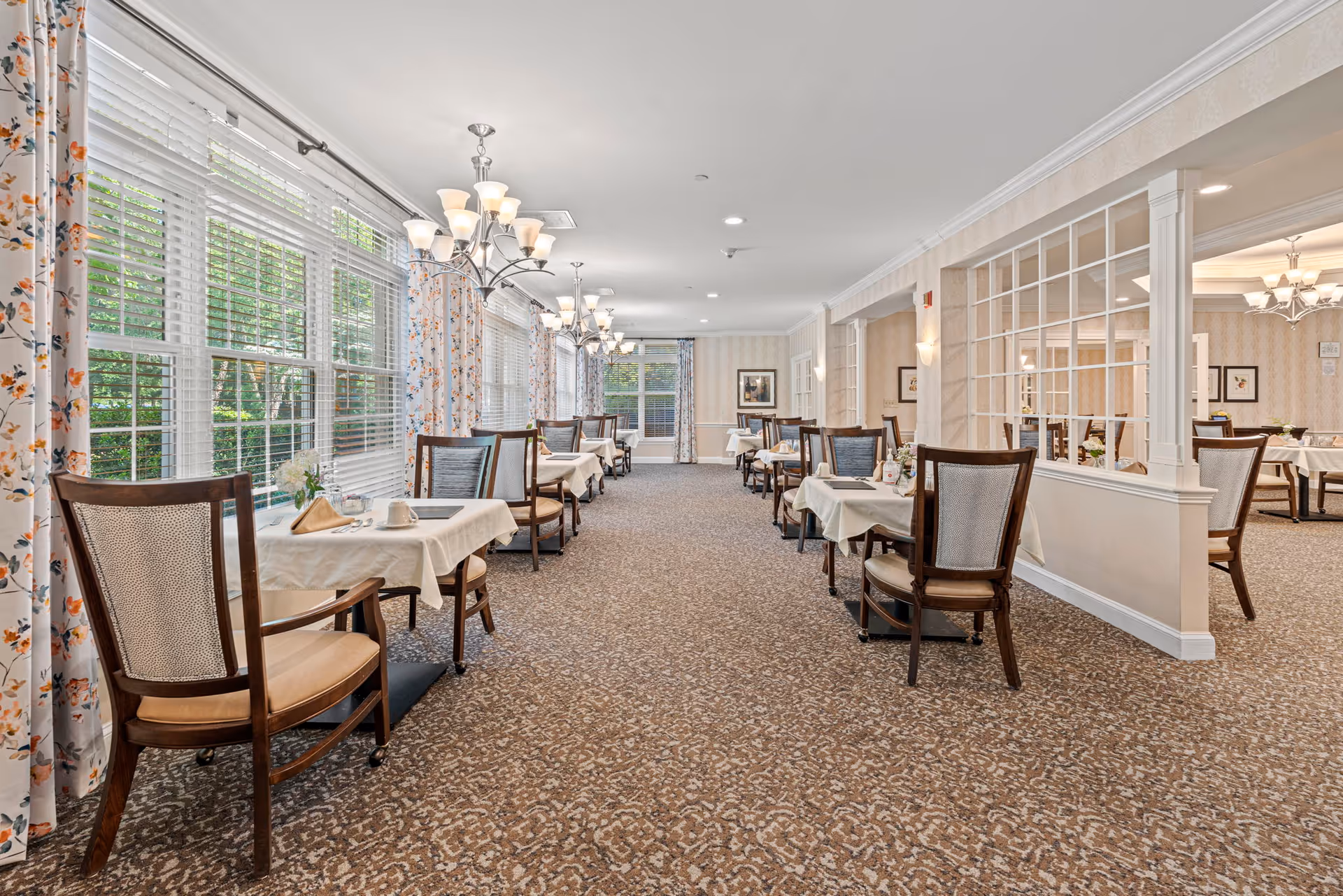 A bright and spacious dining room with multiple tables covered in white tablecloths, each set with cups, napkins, and silverware. The room features large windows with floral curtains allowing natural light to fill the space. Elegant chandeliers hang from the ceiling, and the carpet has a patterned design. There are wooden chairs with cushioned seats arranged around the tables.