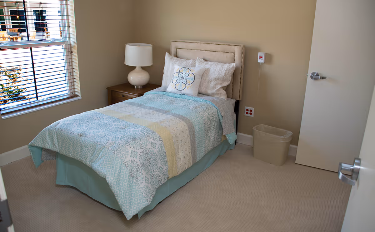 A small, neatly made single bed with a light blue and white patterned comforter and decorative pillows in a simple bedroom. Next to the bed is a wooden nightstand with a white lamp. A window with blinds is on the left wall, and a beige trash bin is on the floor near a closed door on the right.