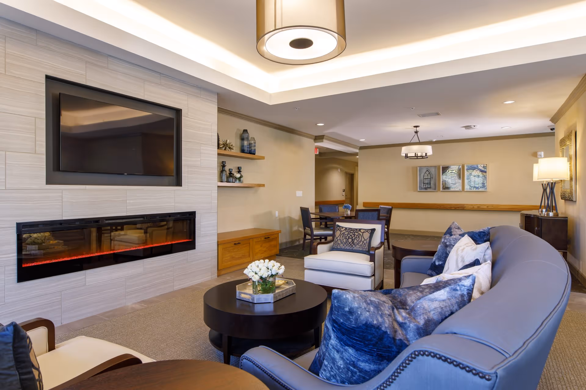 Bright communal lounge with a curved sofa and armchairs around a coffee table, a wall-mounted TV and electric fireplace, and a dining area in the background.