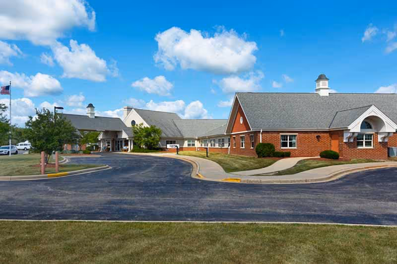 Front exterior of a single-story brick and siding senior care building with a curved driveway and an American flag under a blue sky.