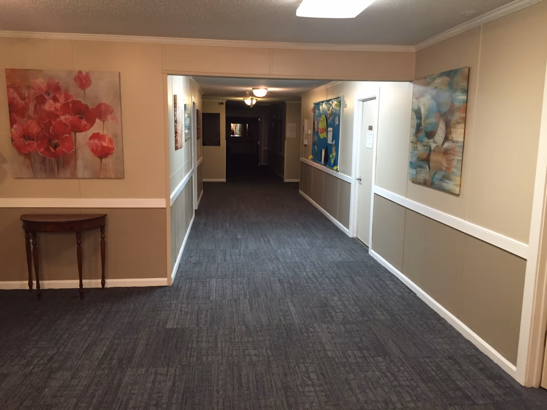 A long indoor hallway with beige walls and blue carpet flooring in an assisted living facility. The hallway is decorated with colorful paintings on the walls and has a small wooden table on the left side. There are several doors along the hallway and ceiling lights providing illumination.