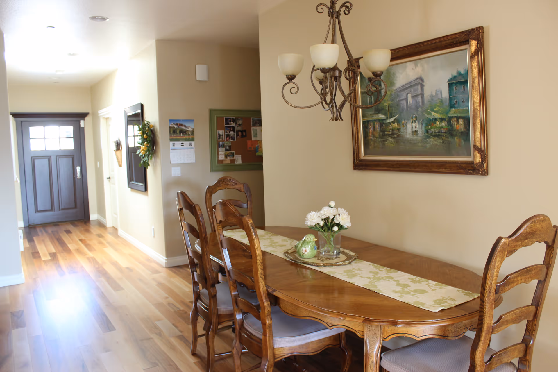 A dining area with a wooden dining table and six matching chairs. The table has a floral table runner and a small vase with white flowers. A chandelier with five lights hangs above the table. On the wall, there is a framed painting depicting a street scene with buildings and trees. The room has wooden flooring and beige walls. In the background, there is a hallway leading to a dark wooden front door with glass panels.