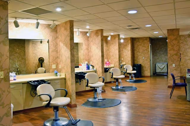 Interior view of a salon area with multiple styling stations. Each station has a large mirror, a beige salon chair, and hair washing sinks. The floor is wooden, and the walls have a patterned wallpaper. The ceiling has recessed lighting and track lights.
