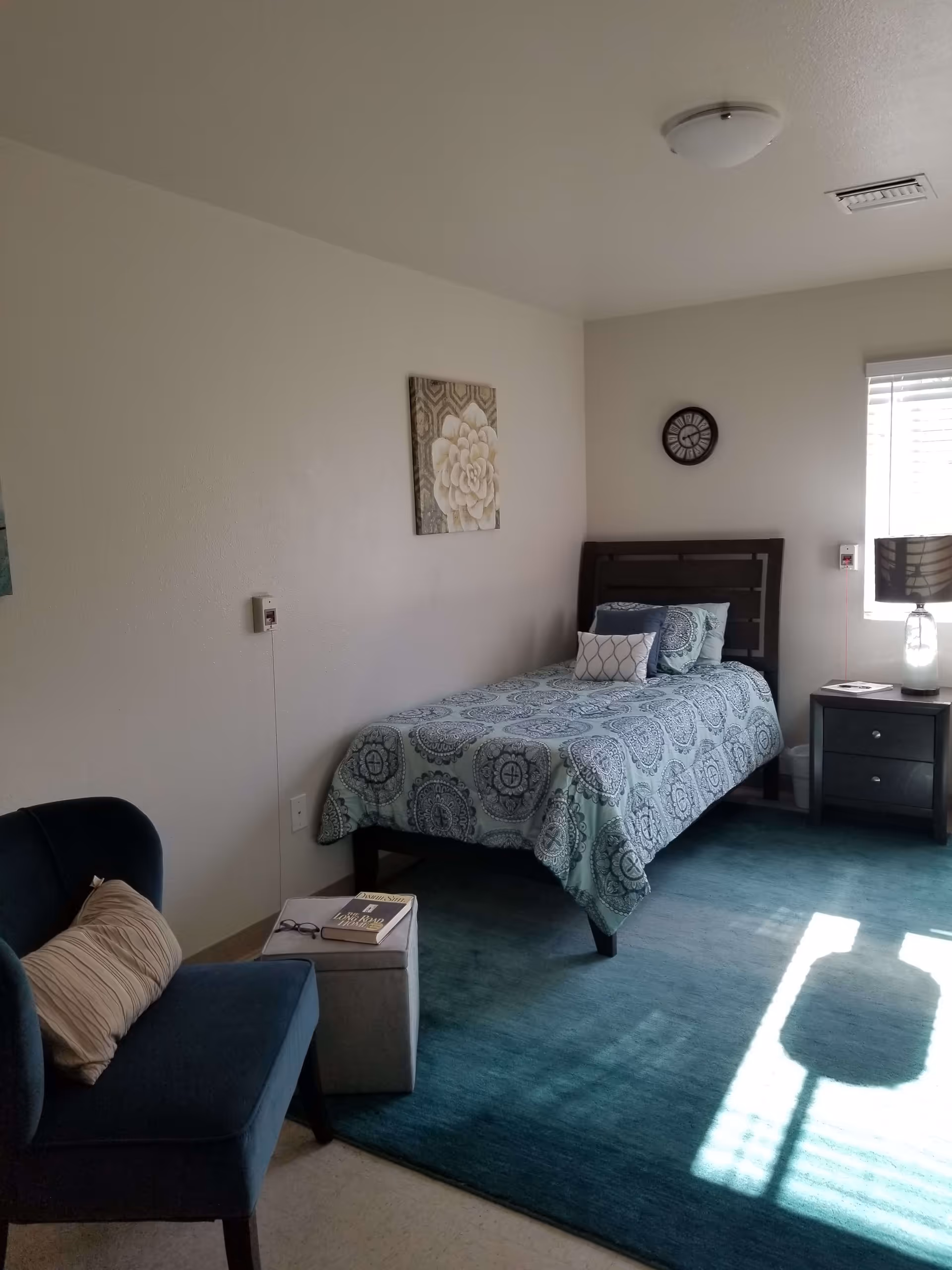 A small, neatly arranged bedroom with a single bed covered in a blue and white patterned bedspread. There is a dark wooden headboard, a nightstand with a lamp, and a clock on the wall above the bed. A blue armchair with a beige pillow sits next to a small ottoman with a book and glasses on it. The room has a large blue rug and a window letting in natural light.