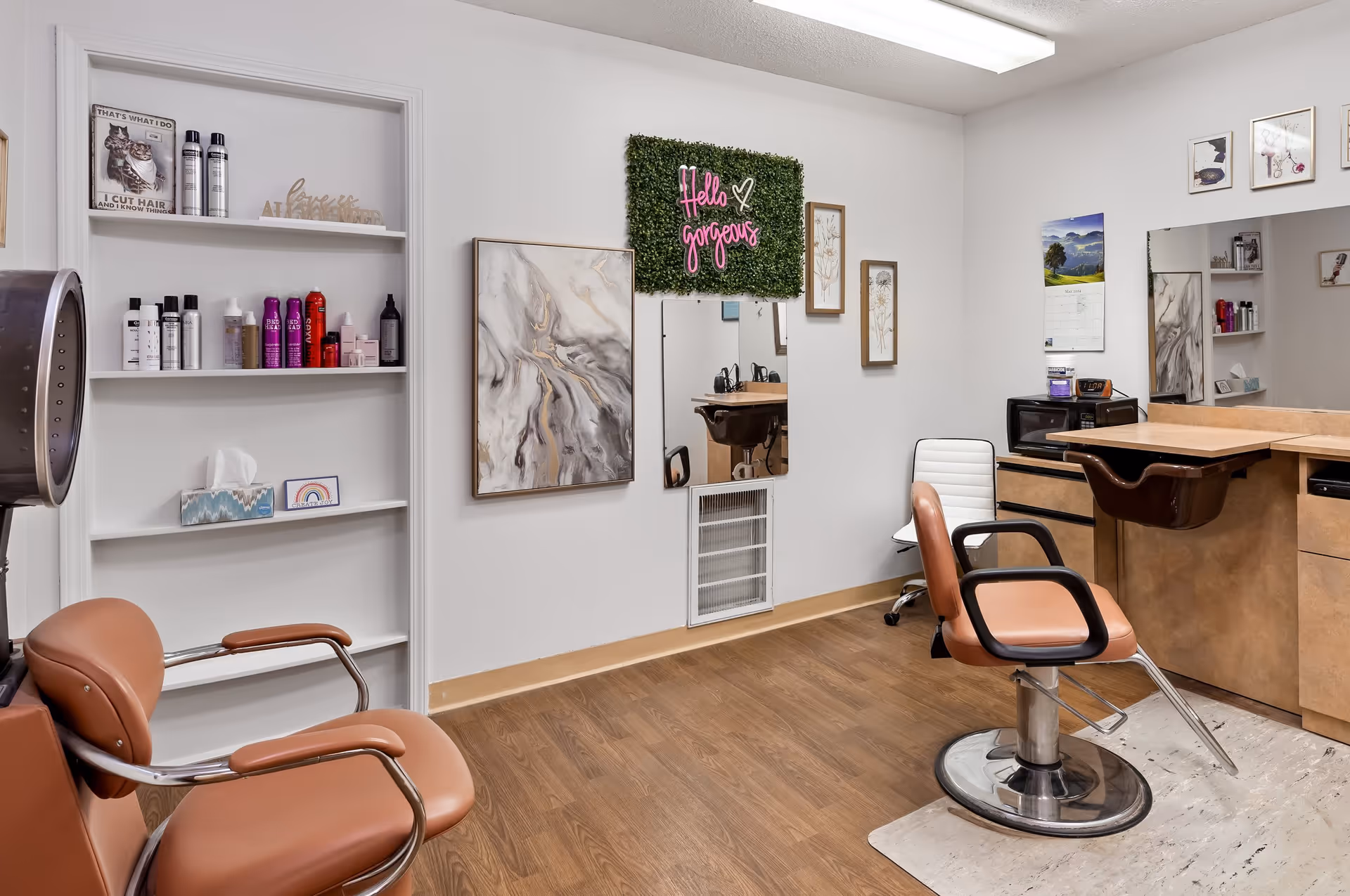Interior of a hair salon with two brown salon chairs, a hair dryer, shelves with hair products, a large mirror, and wall decorations including a neon sign that says 'Hello gorgeous'.