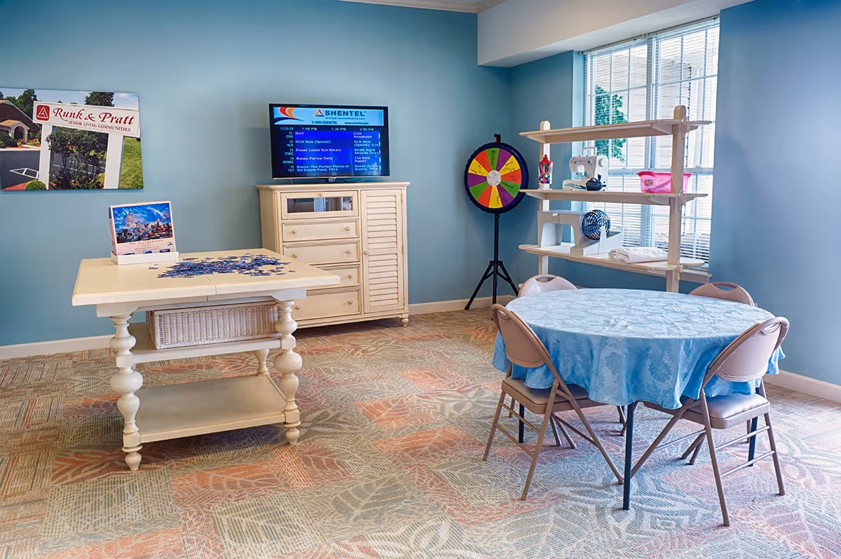 A bright room with light blue walls and patterned carpet featuring a round table covered with a blue tablecloth and four folding chairs. A wooden table with a puzzle in progress is in the foreground. A wooden cabinet with a TV on top is against the wall, and a colorful prize wheel stands next to a shelving unit by a window with blinds. Two framed pictures hang on the wall.