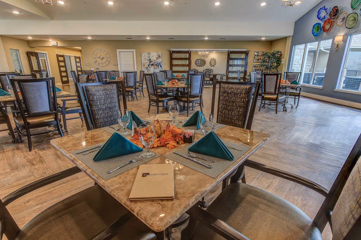 A spacious dining room in a senior living facility named Long Creek, featuring multiple tables set with green folded napkins, glassware, and silverware. The room has wooden flooring, large windows on one side, decorative wall art, and comfortable chairs arranged around the tables.