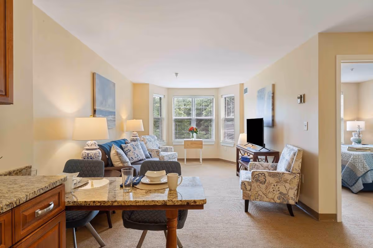 A bright and cozy living room in a senior living facility featuring a granite countertop dining area with two chairs in the foreground, a blue sofa with patterned cushions, two matching armchairs, a TV on a wooden stand, and a bay window with blinds and a small table holding a vase of flowers. A bedroom with a bed and nightstand with a lamp is visible through an open doorway.