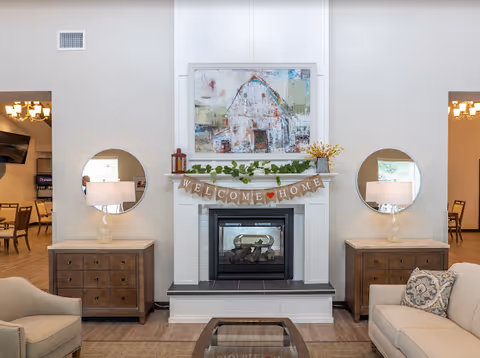 Bright living-room seating area centered on a fireplace with a 'Welcome Home' banner, mirrors, lamps, and artwork.