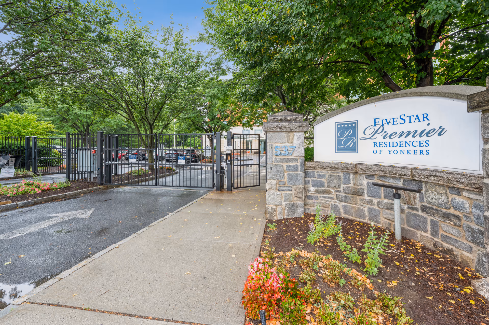 Entrance gate to Five Star Premier Residences of Yonkers with a stone wall sign displaying the facility name. The gate is black metal and closed, surrounded by trees and landscaping with a sidewalk and driveway leading up to it.