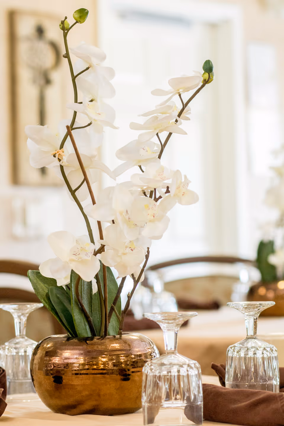 Close-up of a table centerpiece featuring white orchids in a shiny bronze vase, surrounded by upside-down crystal wine glasses and brown napkins in a softly lit dining area.