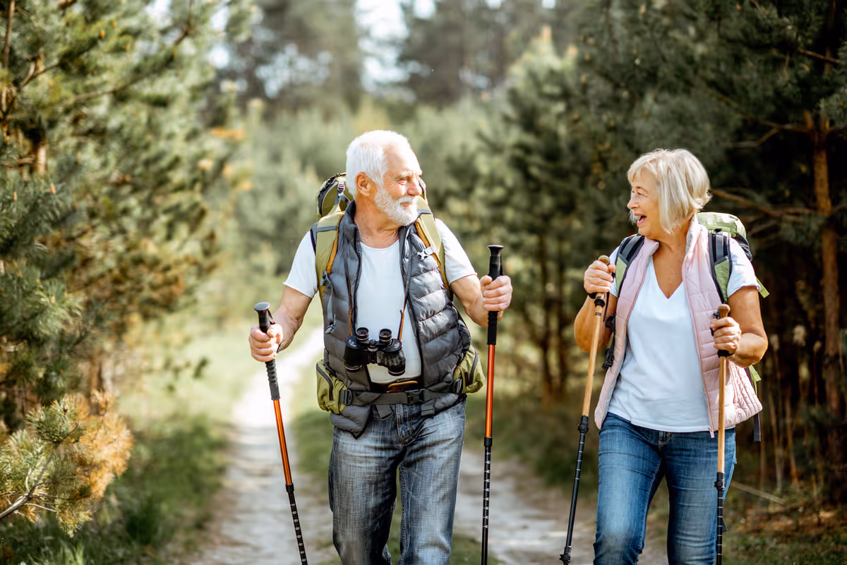 Two older adults hiking on a wooded trail, each carrying a backpack and walking poles.