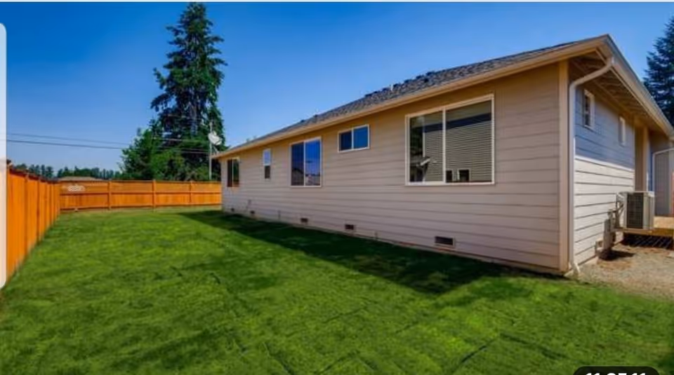 Exterior view of a single-story beige building with several windows, a green lawn, and a wooden fence under a clear blue sky.