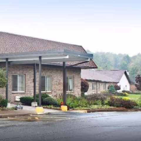 Front view of a single-story brick senior living building with a covered entrance, landscaping, and a wet driveway.