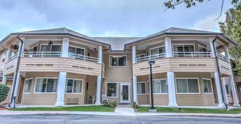 Two-story beige senior living building with balconies and central entrance, across a paved walkway.