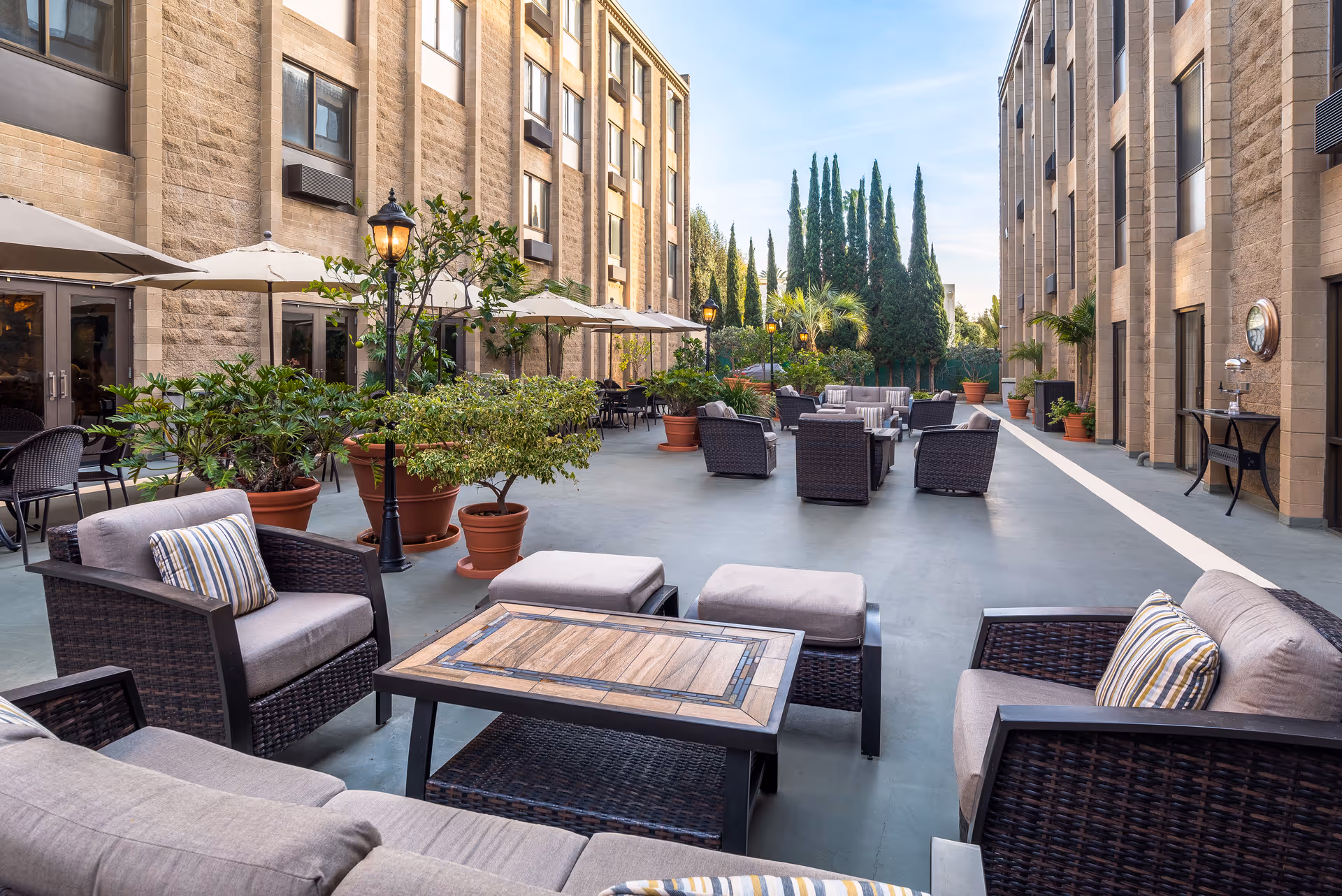 Outdoor courtyard area between two buildings with multiple seating arrangements including cushioned chairs, sofas, and tables. There are potted plants, umbrellas, and lamp posts along the walkway, with tall trees and a clear sky in the background.