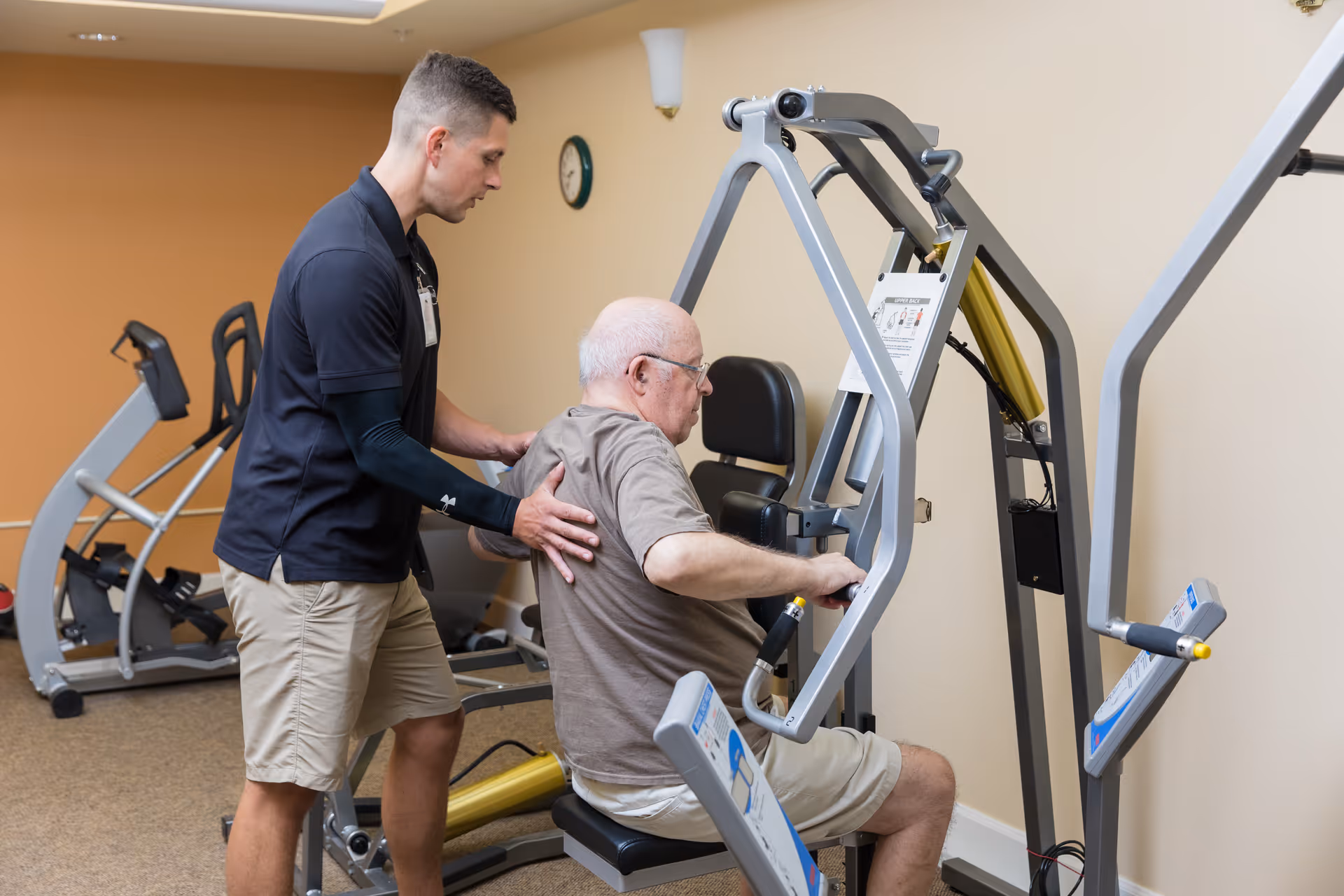 A young male fitness trainer assists an elderly man using a seated weight machine in a gym room with exercise equipment and beige walls.