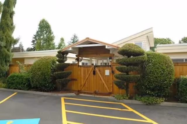 Entrance gate to a facility with a wooden fence and neatly trimmed topiary bushes on either side, surrounded by trees and a parking area with yellow painted lines.