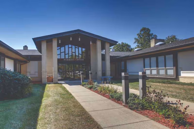 Exterior view of the Odd Fellow-Rebekah Home showing a single-story building with a peaked roof entrance labeled 'Harmony Center'. There is a concrete walkway leading to the entrance, bordered by grass and landscaped plants under a clear blue sky.
