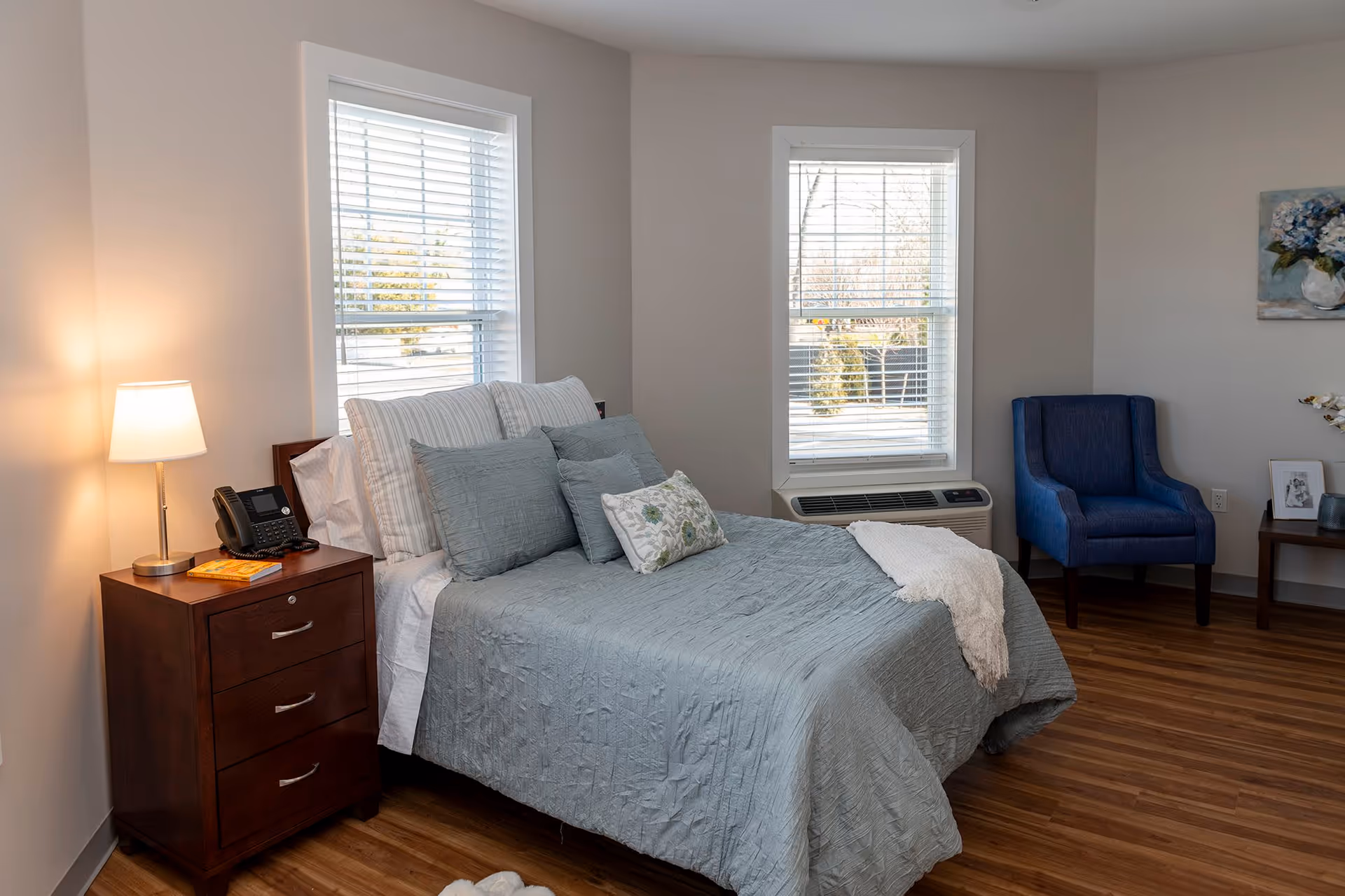 A cozy bedroom with a neatly made bed featuring light blue bedding and multiple pillows. There is a wooden nightstand with a lamp, a telephone, and a book on it. Two windows with white blinds allow natural light into the room. A blue upholstered armchair is placed near the corner, and a small table with decorative items and a framed photo is visible. The room has wooden flooring and light-colored walls.