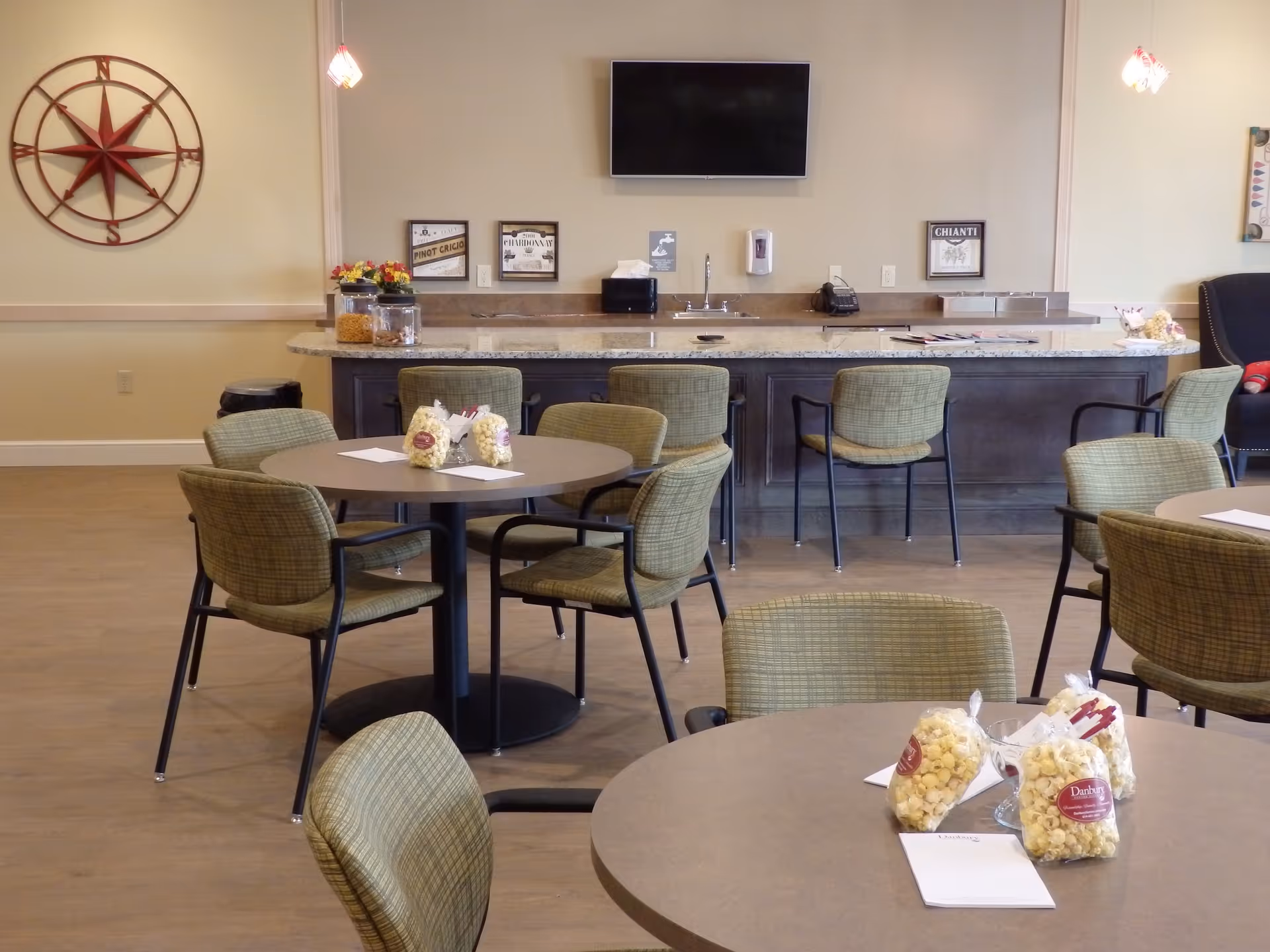 A dining area with round tables and green cushioned chairs. Each table has bags of popcorn and notepads. In the background, there is a counter with bar stools, a sink, a wall-mounted TV, and decorative wall art including a large compass and framed wine labels.