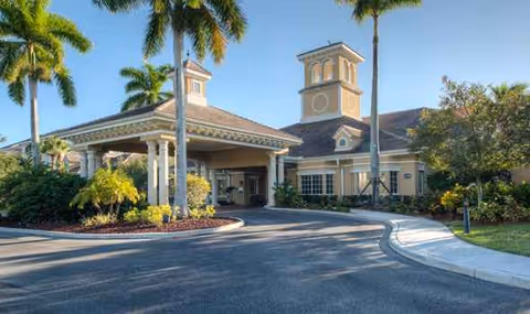 Exterior view of Aston Gardens at Pelican Pointe, featuring porte-cochère entrance and palm trees.