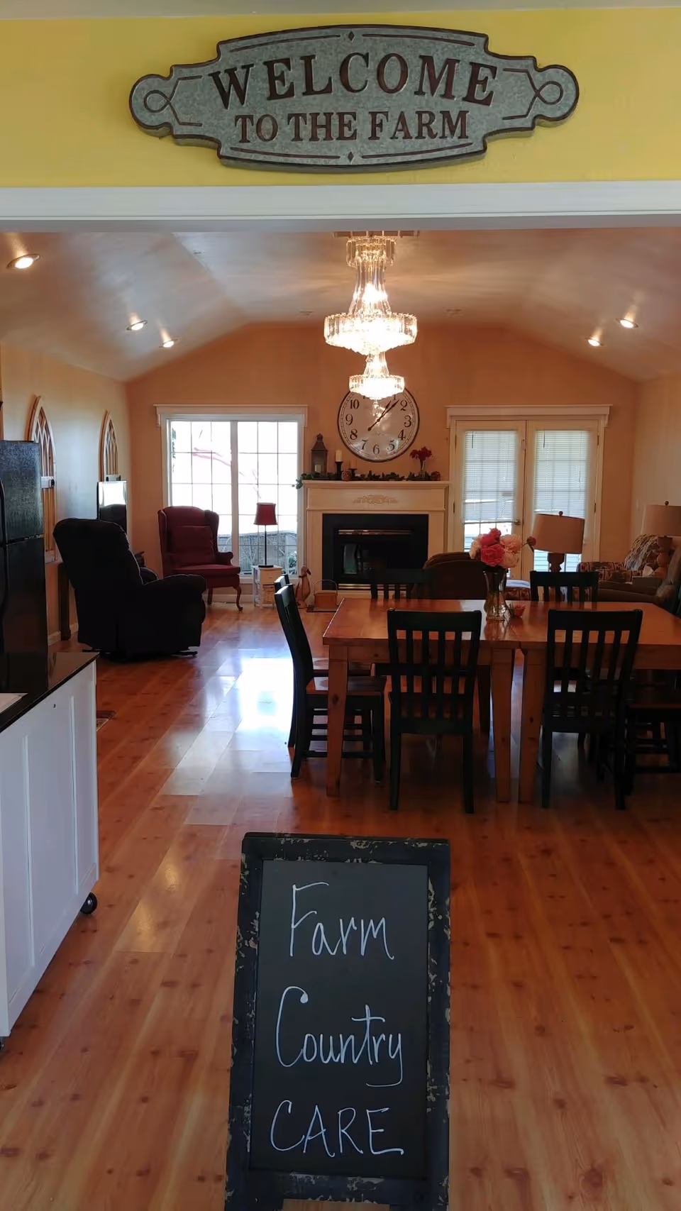 Interior view of a cozy living and dining area with wooden floors, a dining table with chairs, a fireplace with a large clock above it, and comfortable seating including armchairs. A sign above the doorway reads 'WELCOME TO THE FARM' and a chalkboard sign in the foreground says 'Farm Country CARE'.