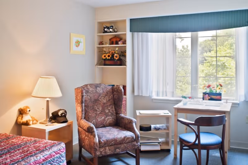 A cozy room with a floral upholstered armchair, a small wooden side table with a lamp, teddy bear, and a hat. There is a bed with a colorful quilt partially visible on the left. A wooden desk with a chair is placed in front of a window with white curtains, and a small shelving unit is next to the desk. The room has soft natural light coming through the window.