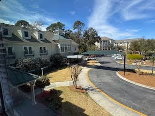 View of the exterior of a senior living facility named Marshes of Skidaway Island, showing a driveway, parking area with cars, landscaped grounds with trees and bushes, and multiple connected buildings under a blue sky with some clouds.