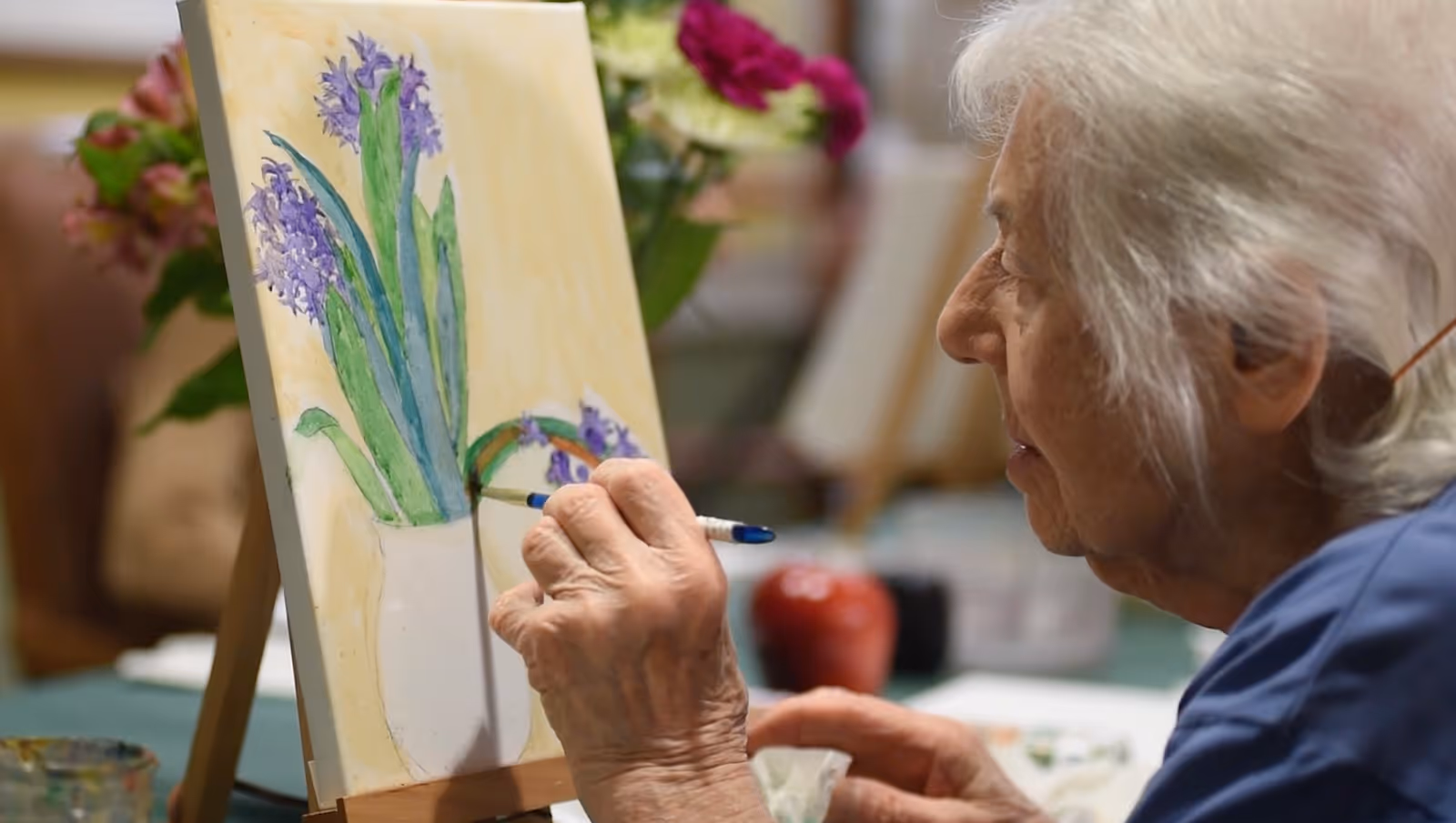 An elderly woman with white hair is painting a picture of purple flowers in a white vase on a canvas set on an easel. The background is softly blurred with other flowers and art supplies visible.