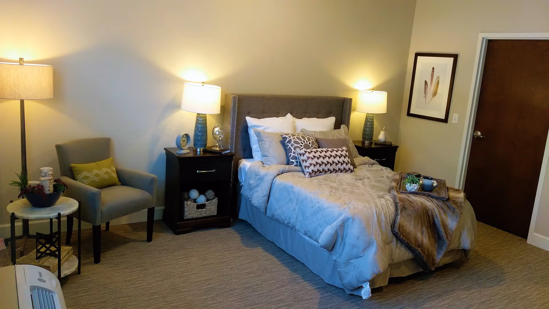 A cozy bedroom with a neatly made bed featuring multiple pillows and a throw blanket. There are two nightstands on either side of the bed, each with a lamp and decorative items. To the left, there is a comfortable armchair with a green cushion and a small round side table with a plant and candle. The room has beige walls, carpeted floor, a framed feather artwork on the wall, and a closed wooden door.