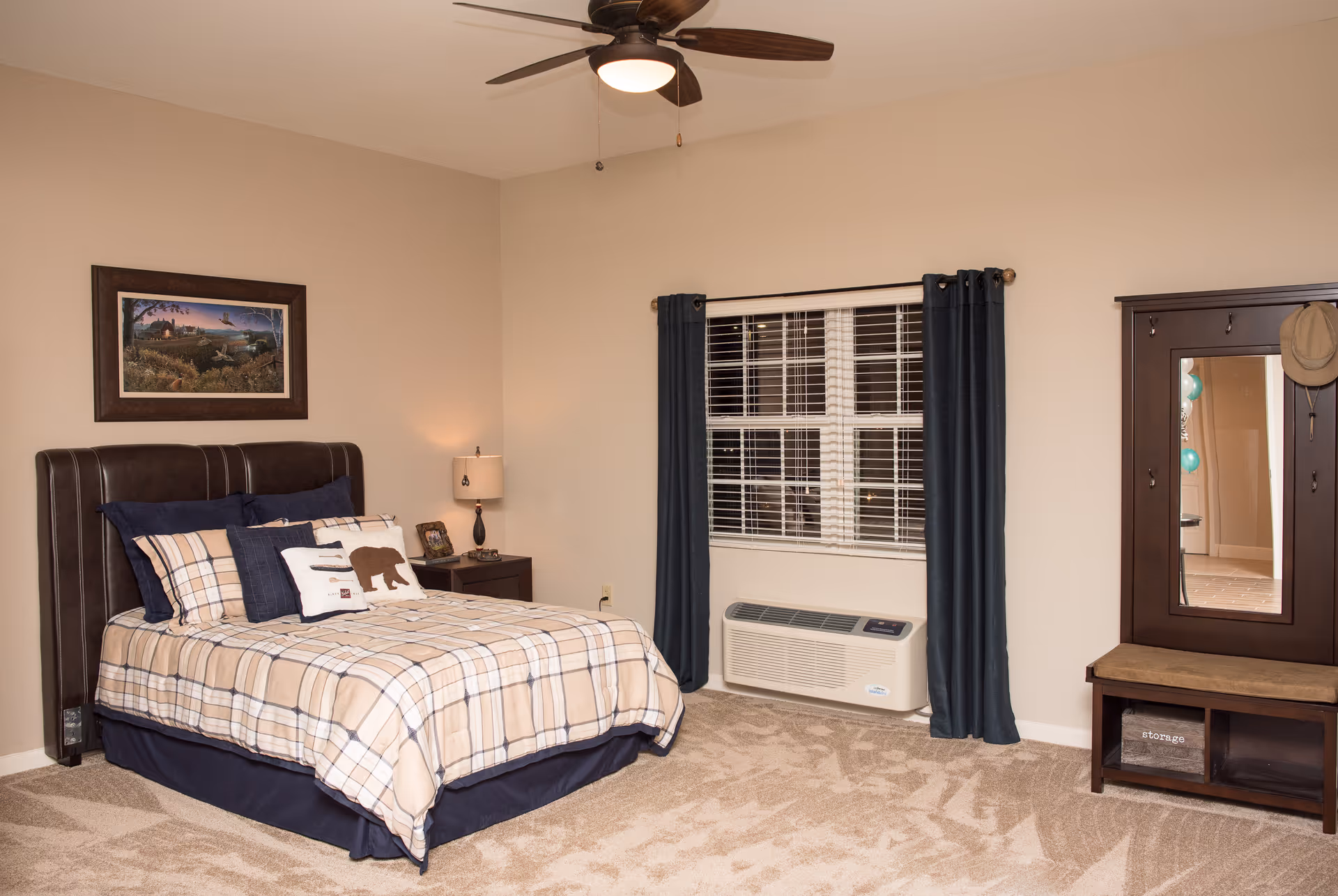 A cozy bedroom with a bed dressed in plaid bedding and multiple pillows, including one with a bear design. There is a nightstand with a lamp and a framed photo next to the bed. A window with dark curtains and blinds is above a wall-mounted air conditioning unit. On the right side, there is a wooden hall tree with a mirror, hooks, a hat hanging, and a cushioned bench with storage compartments underneath. The room has beige walls, carpeted floor, and a ceiling fan with a light.