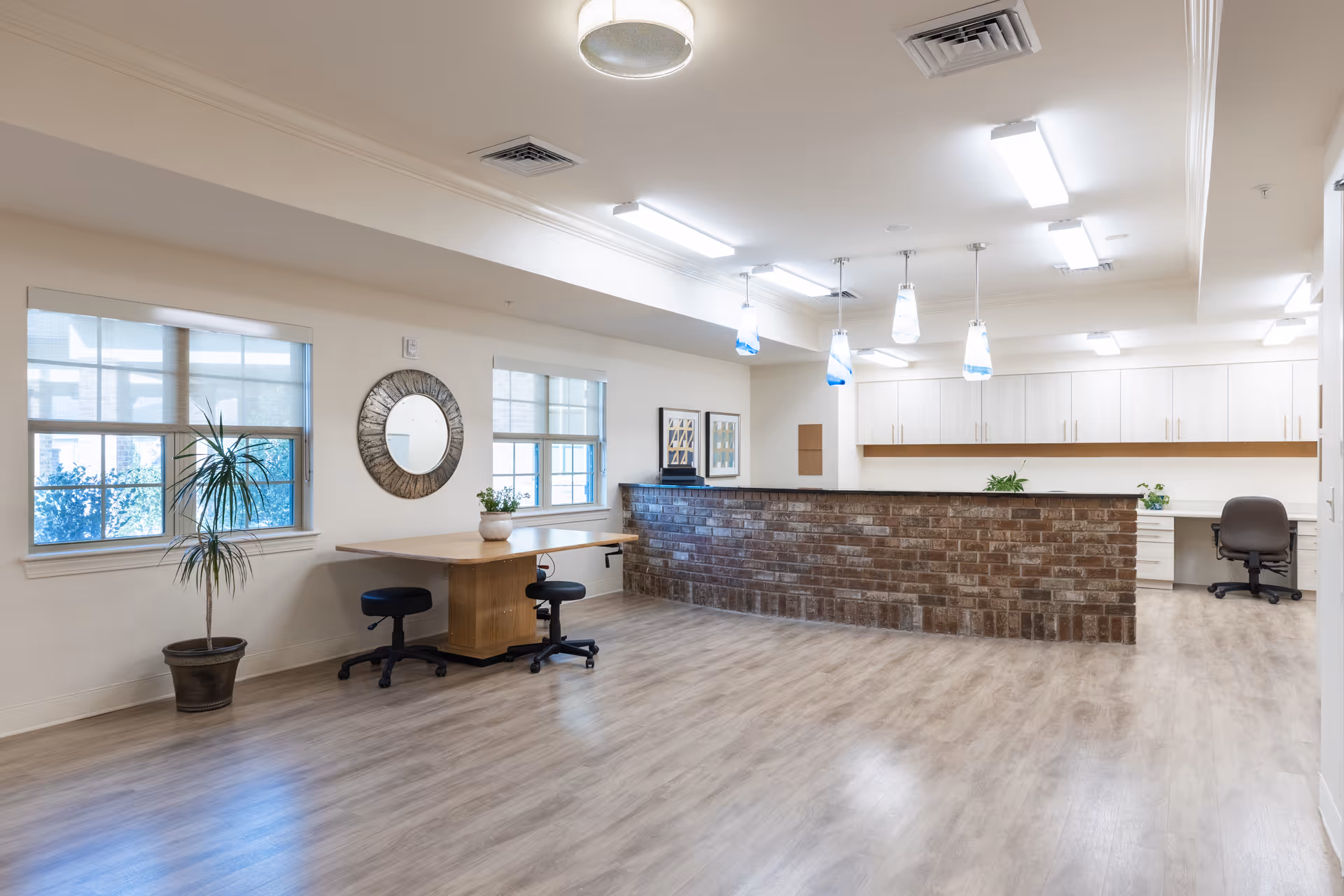 Open bright interior reception area with a brick front desk, a wooden table with stools, windows, and office chairs.